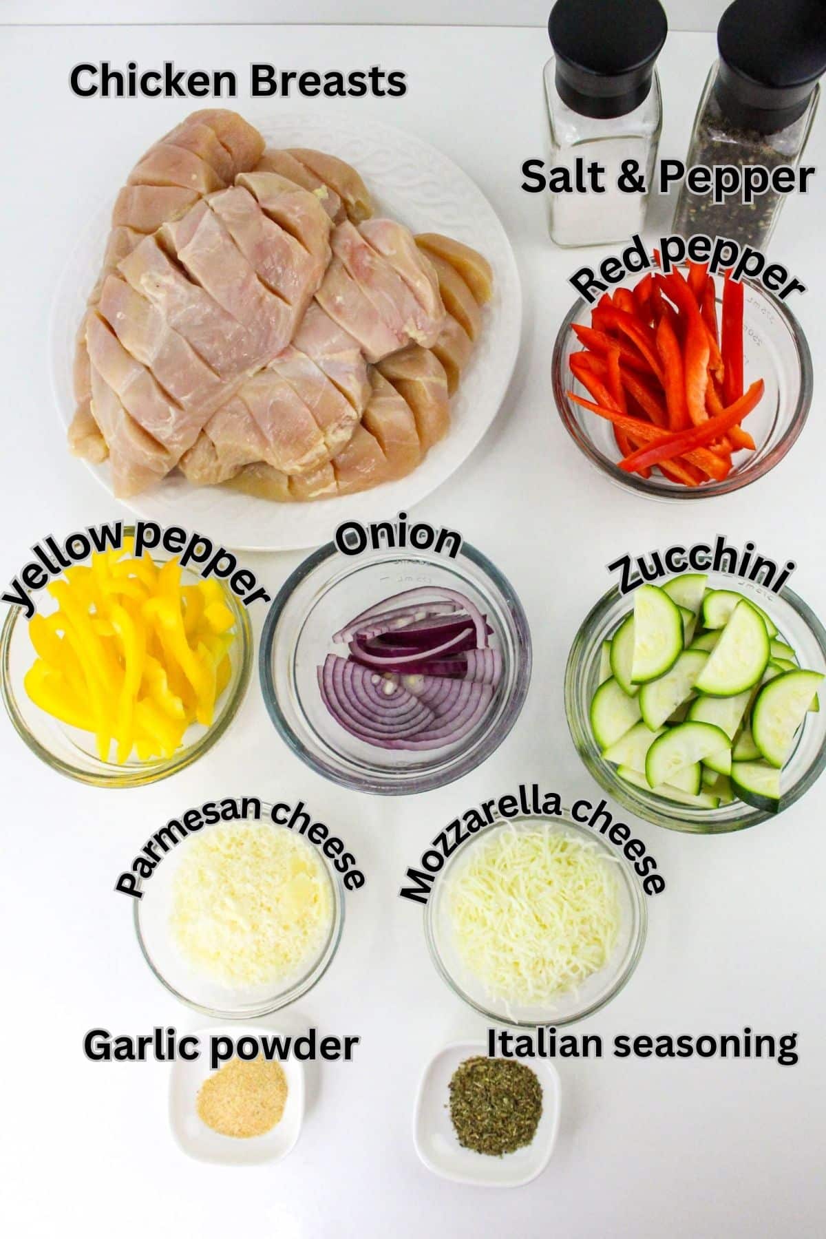 Ingredients on a table: raw chicken breasts, salt, pepper, sliced red and yellow peppers, zucchini, onion, grated Parmesan and mozzarella cheese, garlic powder, and Italian seasoning.