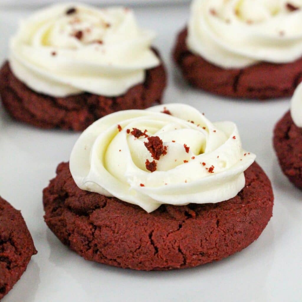 Red velvet cookies topped with swirls of white frosting and sprinkled with red crumbs are displayed on a white surface.