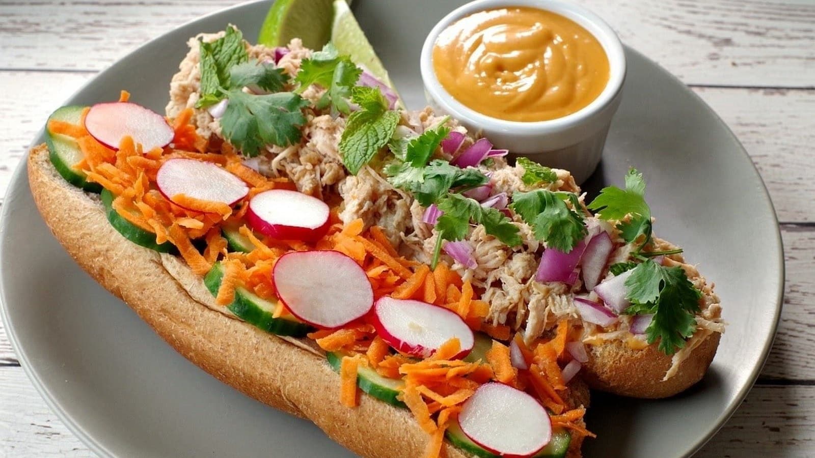 A sandwich with shredded chicken, radishes, cucumbers, carrots, cilantro, and purple onions on a gray plate, next to a small bowl of dipping sauce.