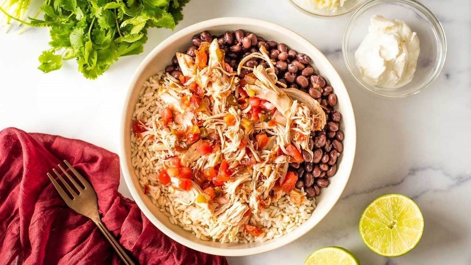 A bowl of rice topped with shredded chicken, black beans, and diced tomatoes, surrounded by fresh cilantro, lime halves, sour cream, and a red napkin with a fork on a white surface.