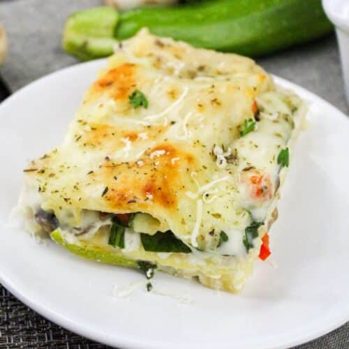 A slice of vegetable lasagna topped with melted cheese and herbs on a white plate.