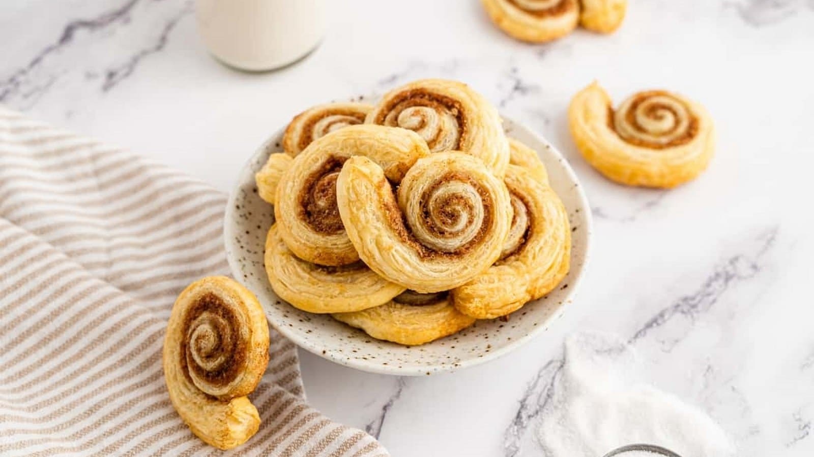 A bowl of cinnamon swirl pastries on a marble surface, with additional pastries, a striped cloth, and a glass of milk nearby.