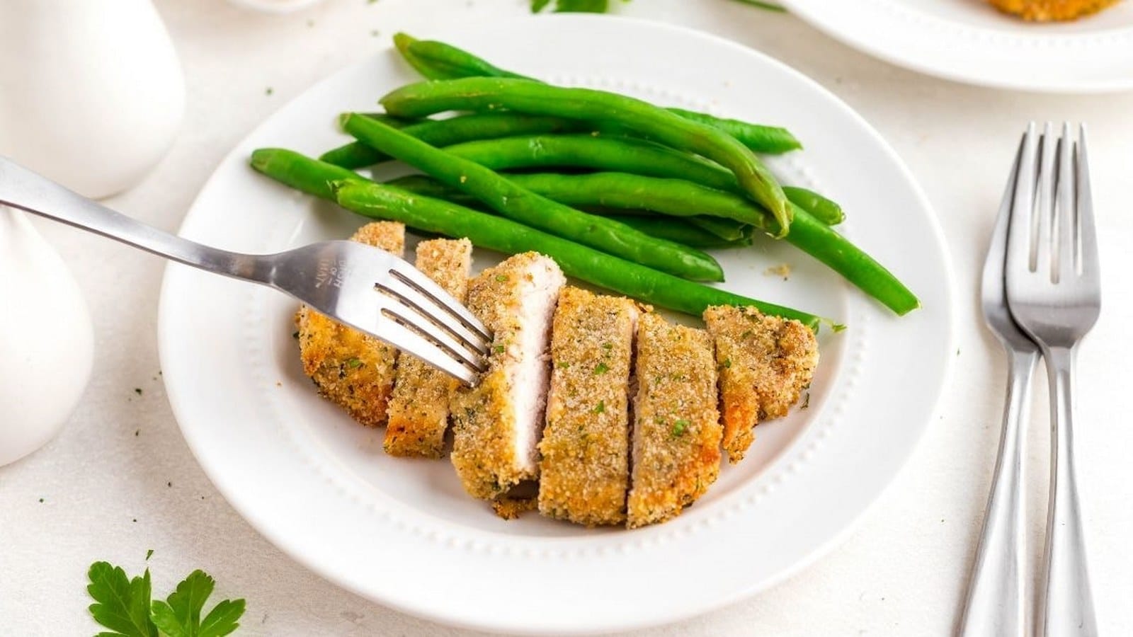 A fork holds a piece of sliced breaded chicken on a plate with green beans. Additional forks and plates are visible in the background.