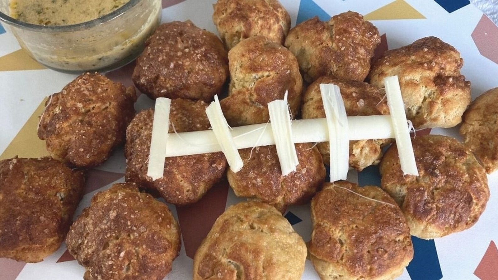 A platter of round, brown snacks resembling a football, with a string cheese decoration, and a small bowl of dipping sauce on a geometric-patterned tablecloth.