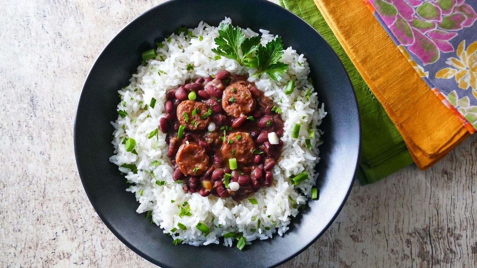 A black bowl of rice topped with red beans, sausage slices, and green onions, garnished with parsley. It is set on a wooden surface with colorful napkins beside it.