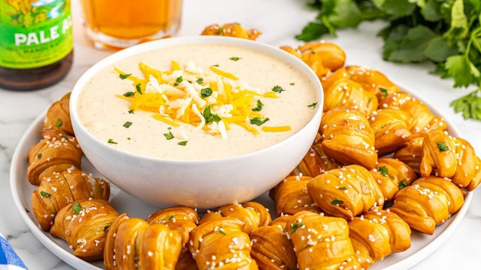 A bowl of cheese dip garnished with shredded cheese and herbs is surrounded by pretzel bites on a plate. A beer bottle, a glass, and fresh parsley are in the background.