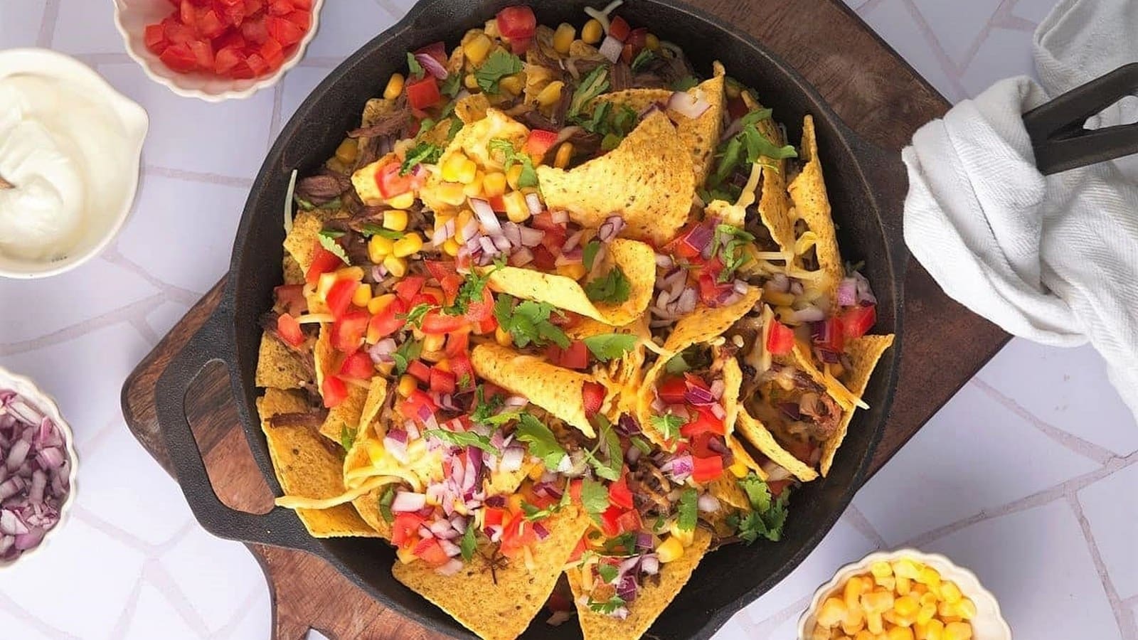 A skillet of nachos topped with cheese, diced tomatoes, onions, corn, and cilantro, surrounded by small bowls of sour cream, chopped onions, and corn.