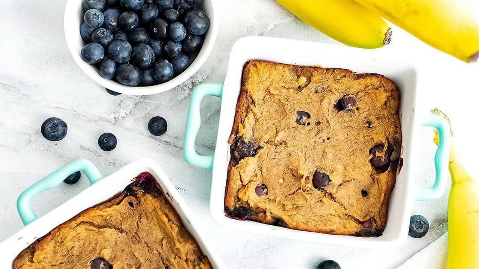 Two square dishes of baked banana blueberry oatmeal on a tabletop, with bananas and a bowl of blueberries beside them.