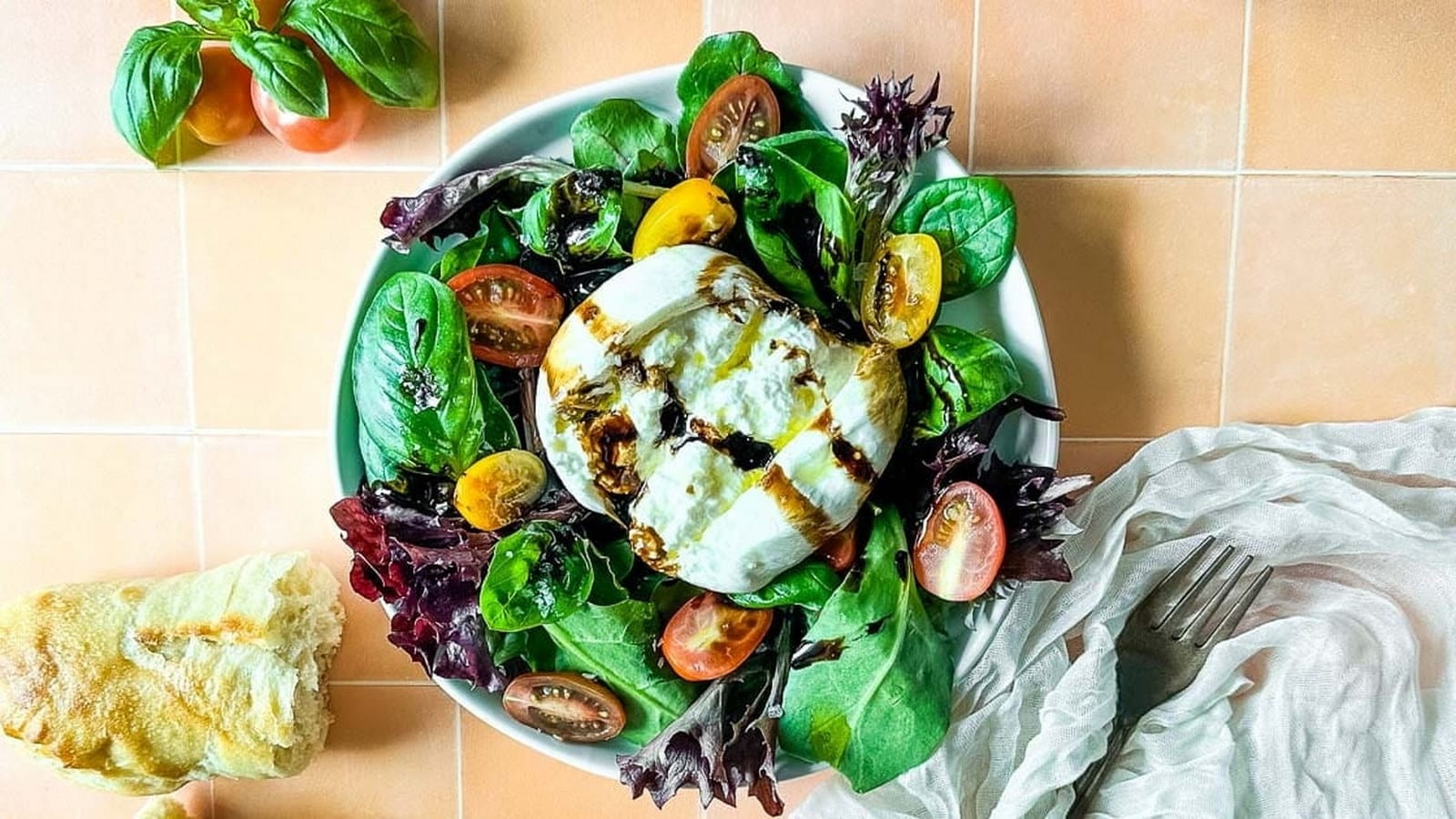 A fresh salad with mixed greens, cherry tomatoes, and a ball of burrata drizzled with balsamic glaze. A piece of bread and basil leaves are on the side.