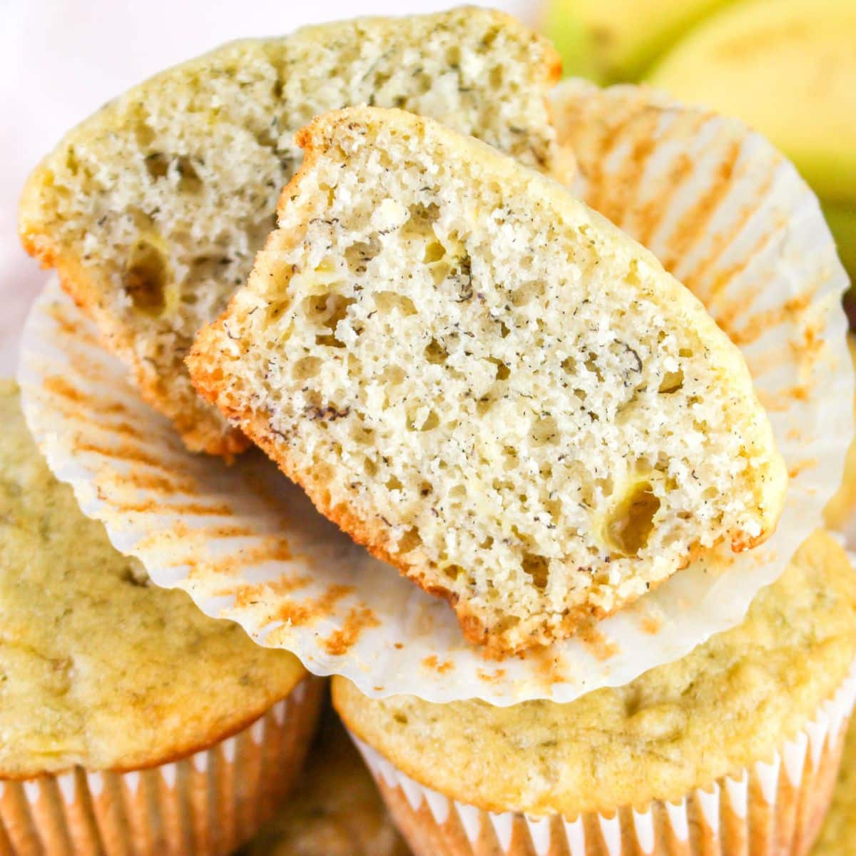 Sliced banana muffin on a cupcake liner, showing a moist, speckled interior, with whole muffins in the background.