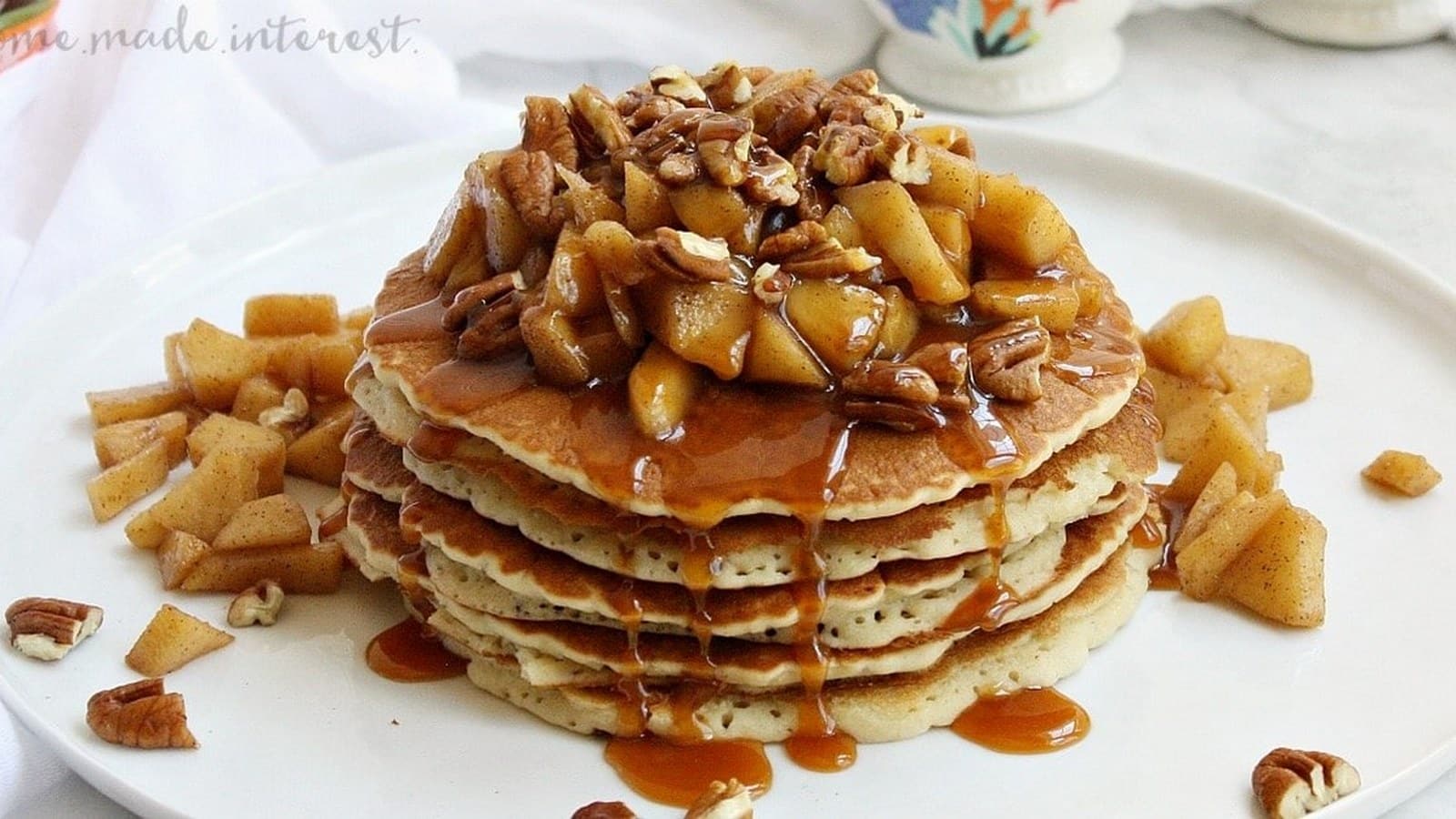 Stack of pancakes topped with diced apples, pecans, and caramel sauce on a white plate.