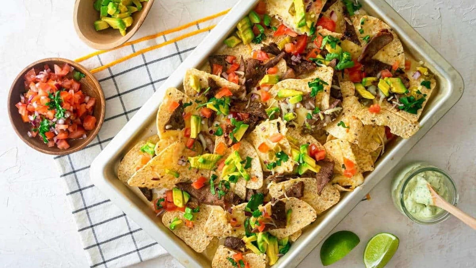 A tray of loaded nachos topped with sliced beef, avocado, diced tomatoes, jalapeños, and cilantro. Accompanied by bowls of salsa and guacamole, and lime wedges on the side.