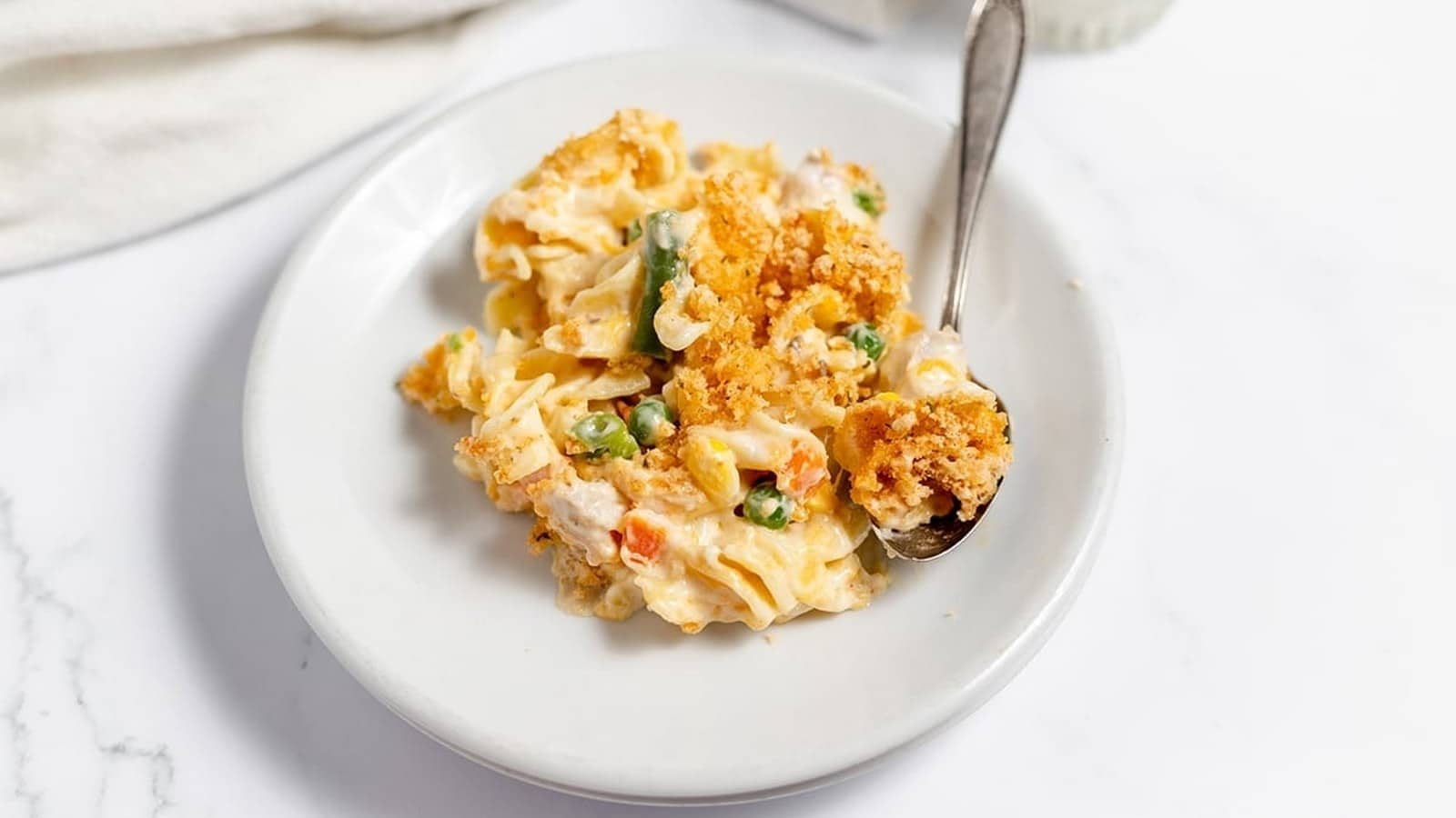 A plate of creamy pasta casserole with peas, carrots, and a golden breadcrumb topping, accompanied by a spoon.