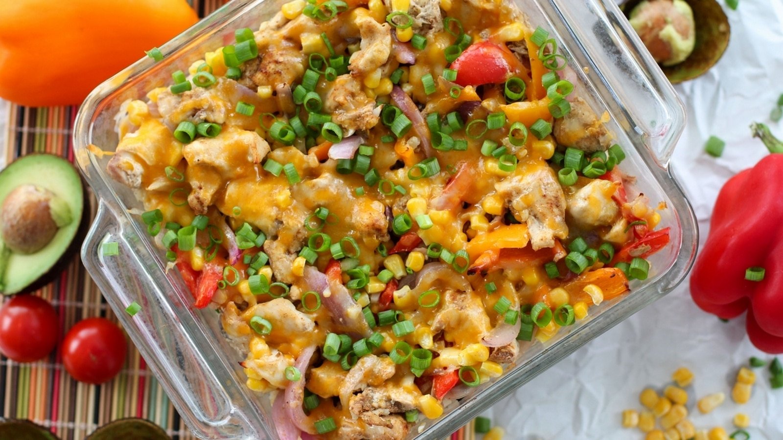 A baked casserole dish filled with chicken, melted cheese, green onions, corn, and diced red and yellow bell peppers, surrounded by avocados, tomatoes, and whole bell peppers.