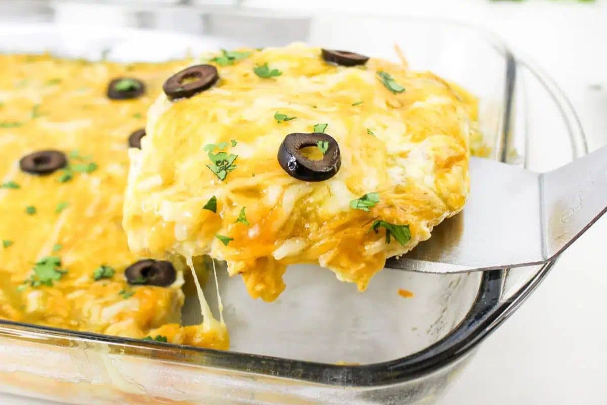 A spatula lifting a slice of cheesy casserole topped with black olives and herbs from a glass baking dish.