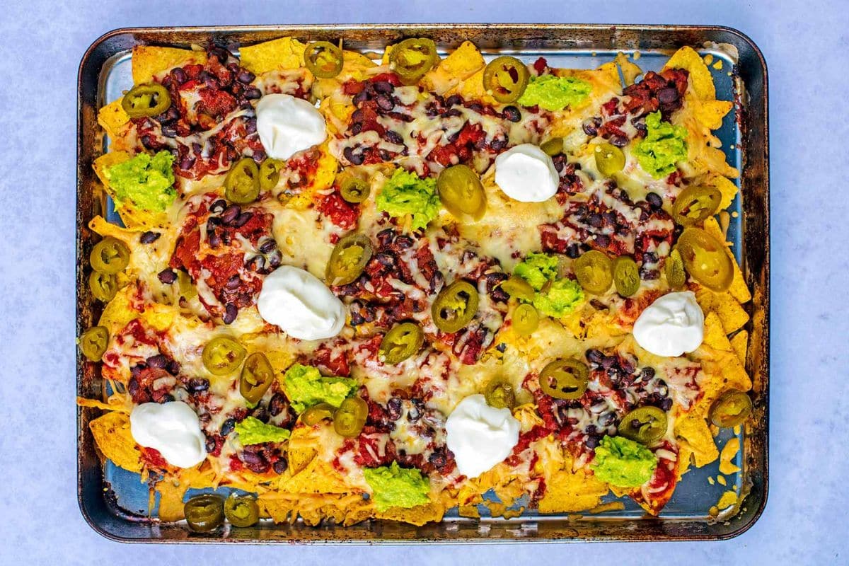 Baked nachos on a tray topped with melted cheese, jalapeños, black beans, guacamole, salsa, and sour cream.