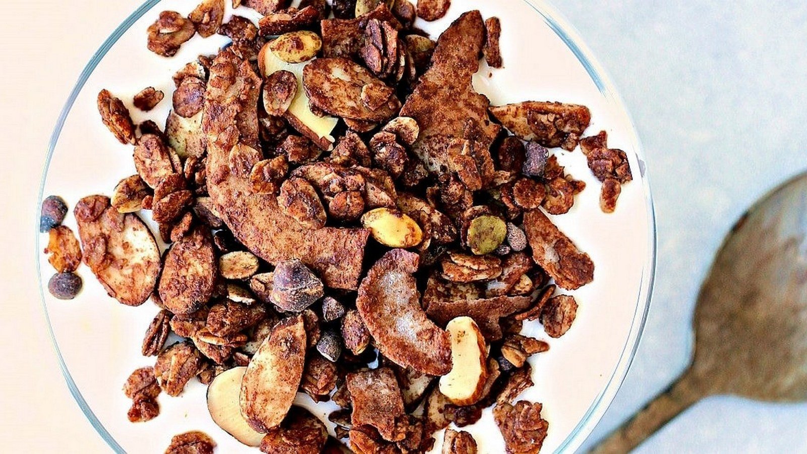 A bowl of yogurt topped with granola, almonds, and other nuts sits beside a wooden spoon on a white surface.