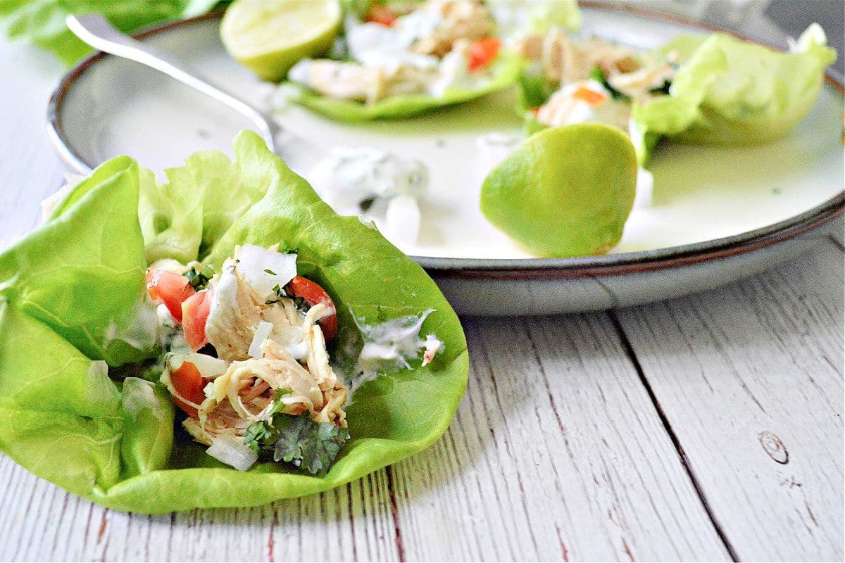 Lettuce wraps with shredded chicken, tomatoes, and herbs on a plate, accompanied by lime halves.