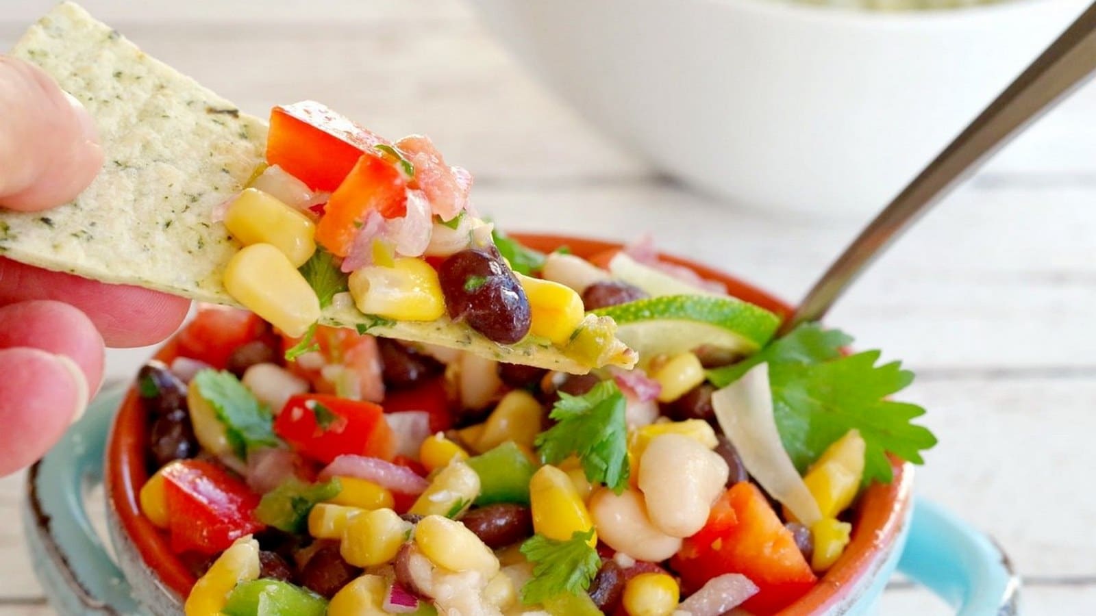 A hand holds a chip topped with a mix of corn, black beans, tomatoes, onions, and cilantro above a bowl of salad. A metal spoon is in the bowl.