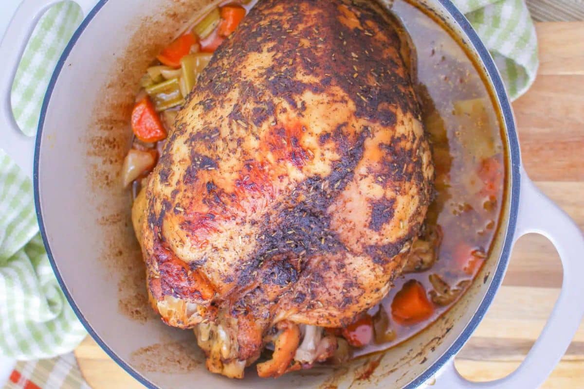Roast chicken in a pot with carrots, onions, and celery on a wooden surface.