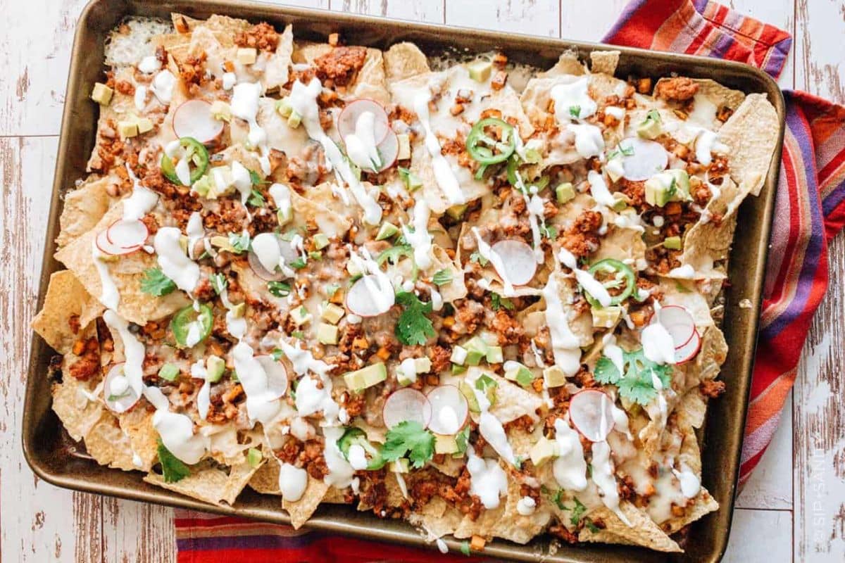 A baking sheet filled with loaded nachos topped with ground meat, melted cheese, radishes, jalapeños, sour cream, and cilantro on a striped cloth.