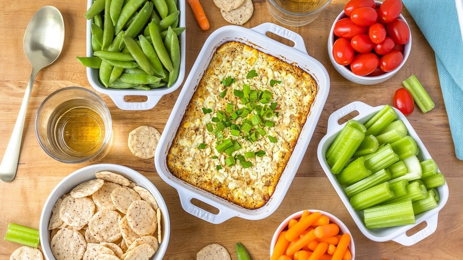 A white baking dish with baked dip, surrounded by bowls of cherry tomatoes, snap peas, celery, crackers, carrot sticks, and glasses of a light beverage on a wooden table.