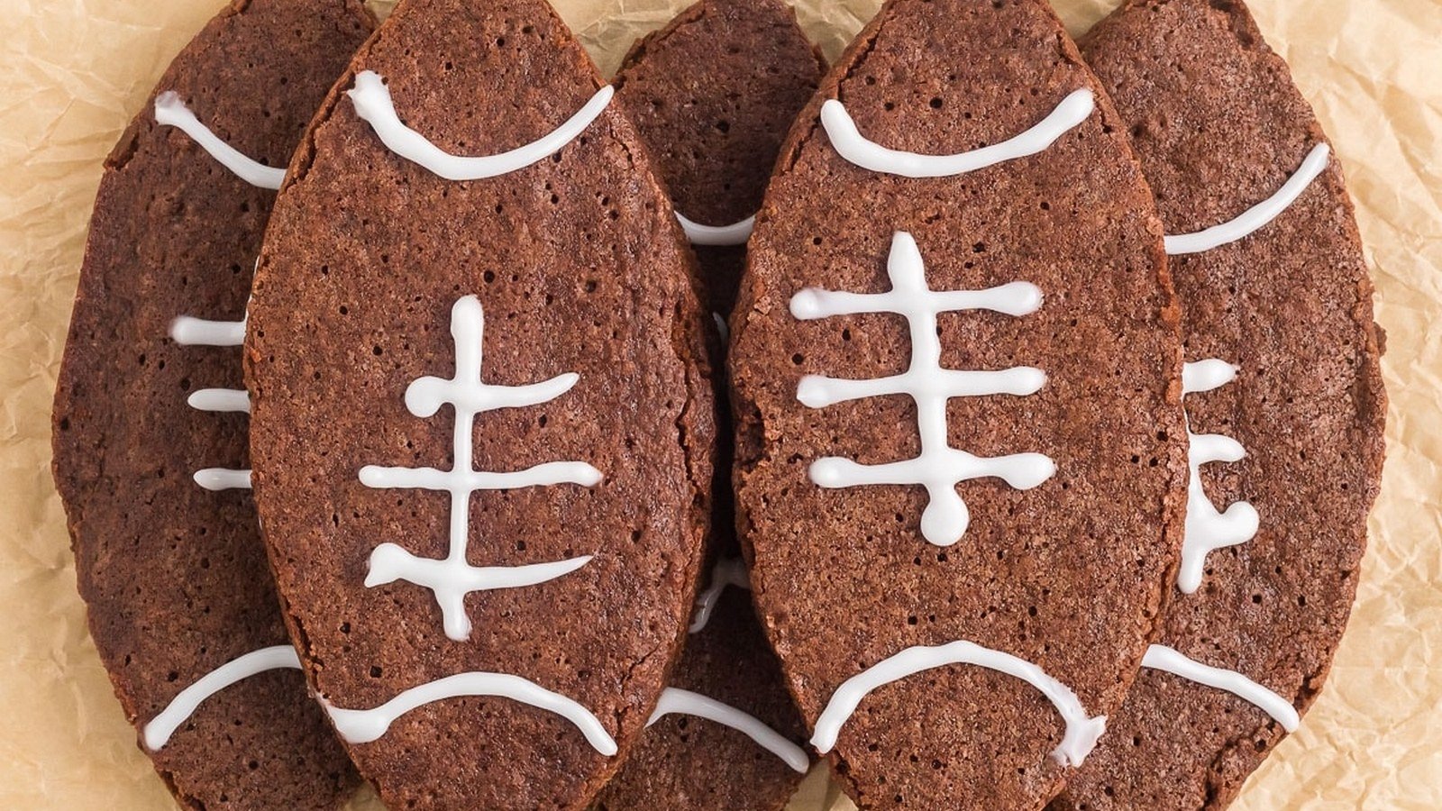 Five chocolate cookies shaped like footballs, decorated with white icing to resemble laces, arranged on parchment paper.