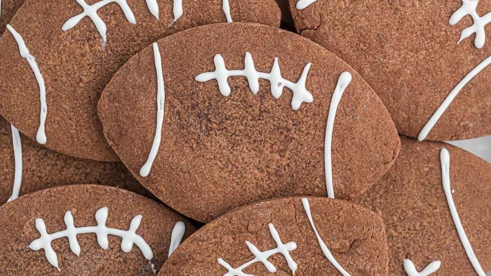 Brown football-shaped cookies with white icing resembling stitches are stacked together.