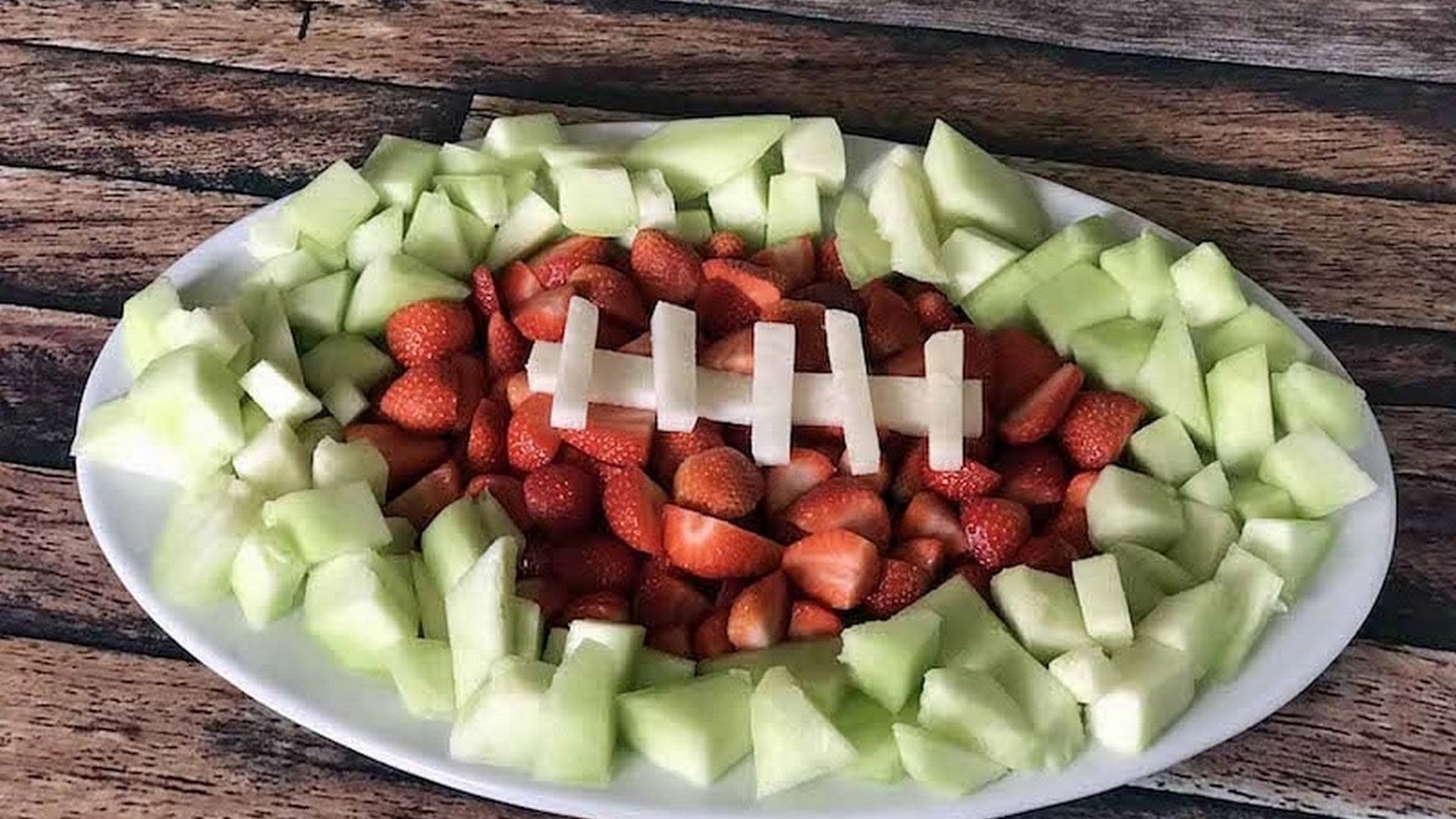 A platter of strawberries and melon arranged in the shape of a football on a wooden table.