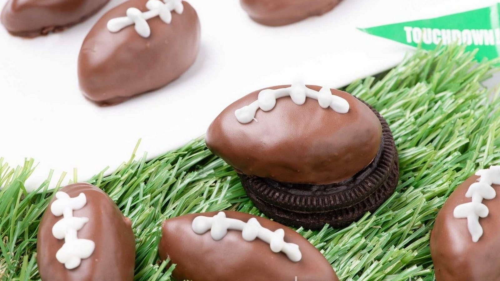 Chocolate-covered Oreos shaped like footballs, decorated with white icing, are displayed on artificial grass.