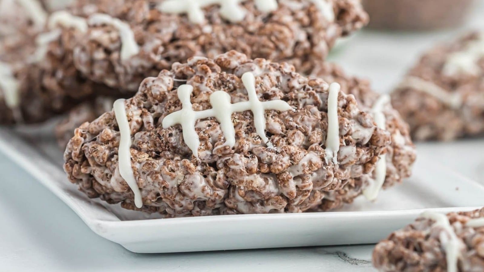 Chocolate rice cereal treats shaped like footballs, decorated with white icing to resemble stitching, placed on a white plate.