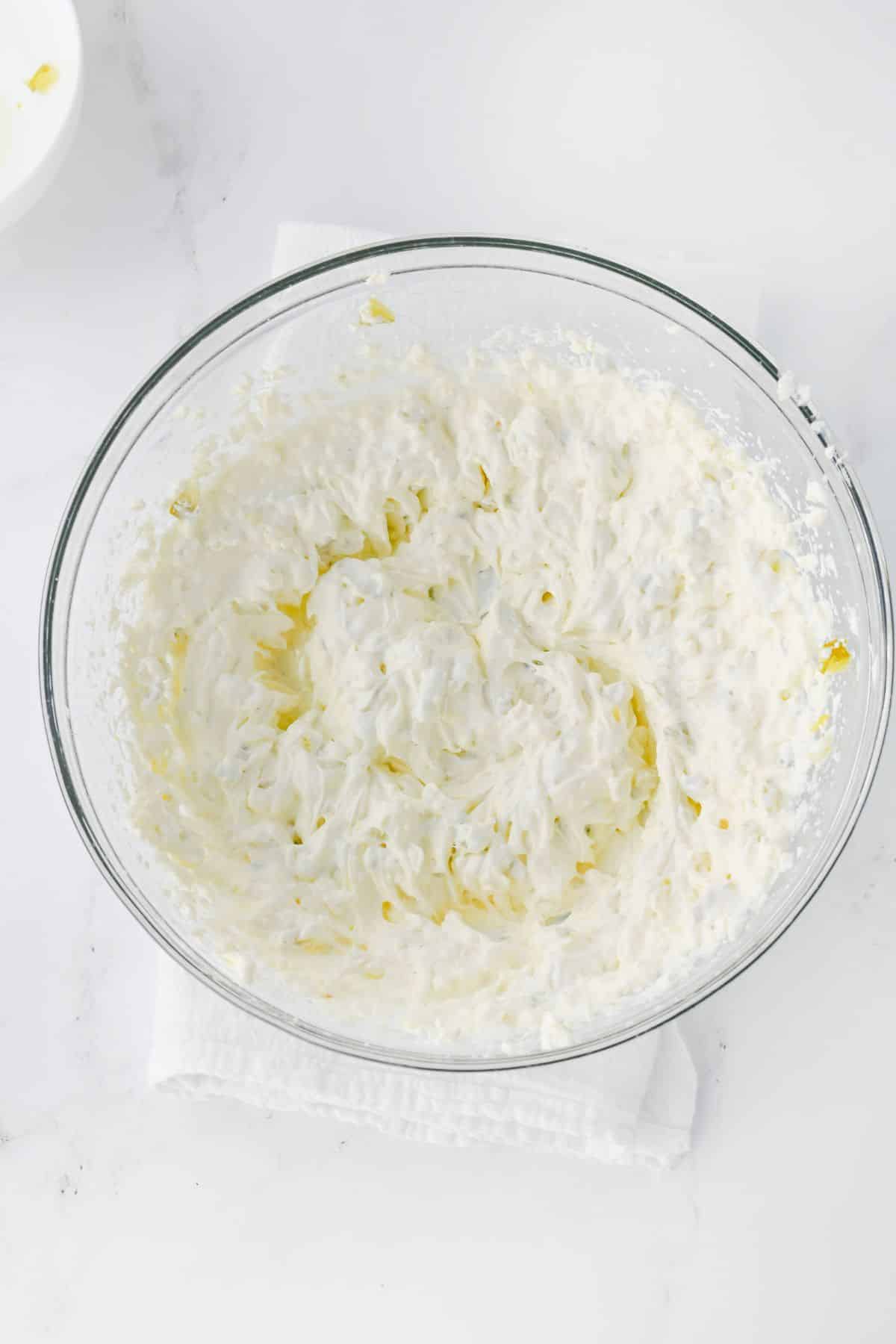 A glass bowl containing a thick, creamy, white mixture, likely a whipped dessert or topping, placed on a white cloth on a countertop.