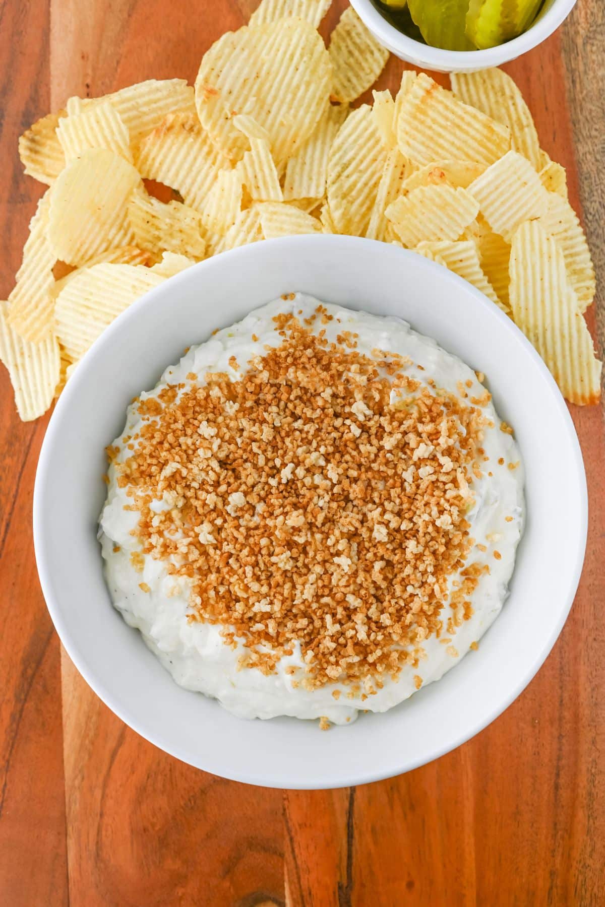 A bowl of creamy dip topped with breadcrumbs, surrounded by crinkle-cut potato chips on a wooden surface.