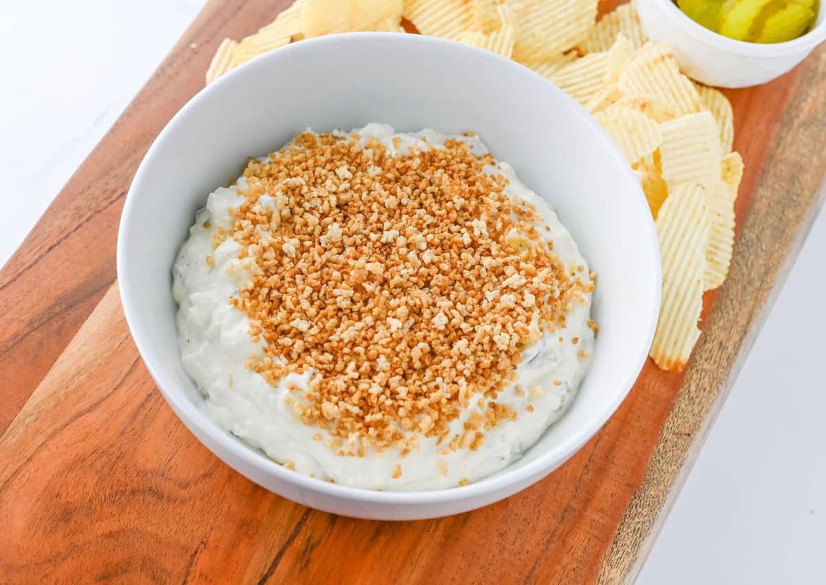 A bowl of creamy dip topped with breadcrumbs, placed on a wooden board with potato chips and a small bowl of pickles.