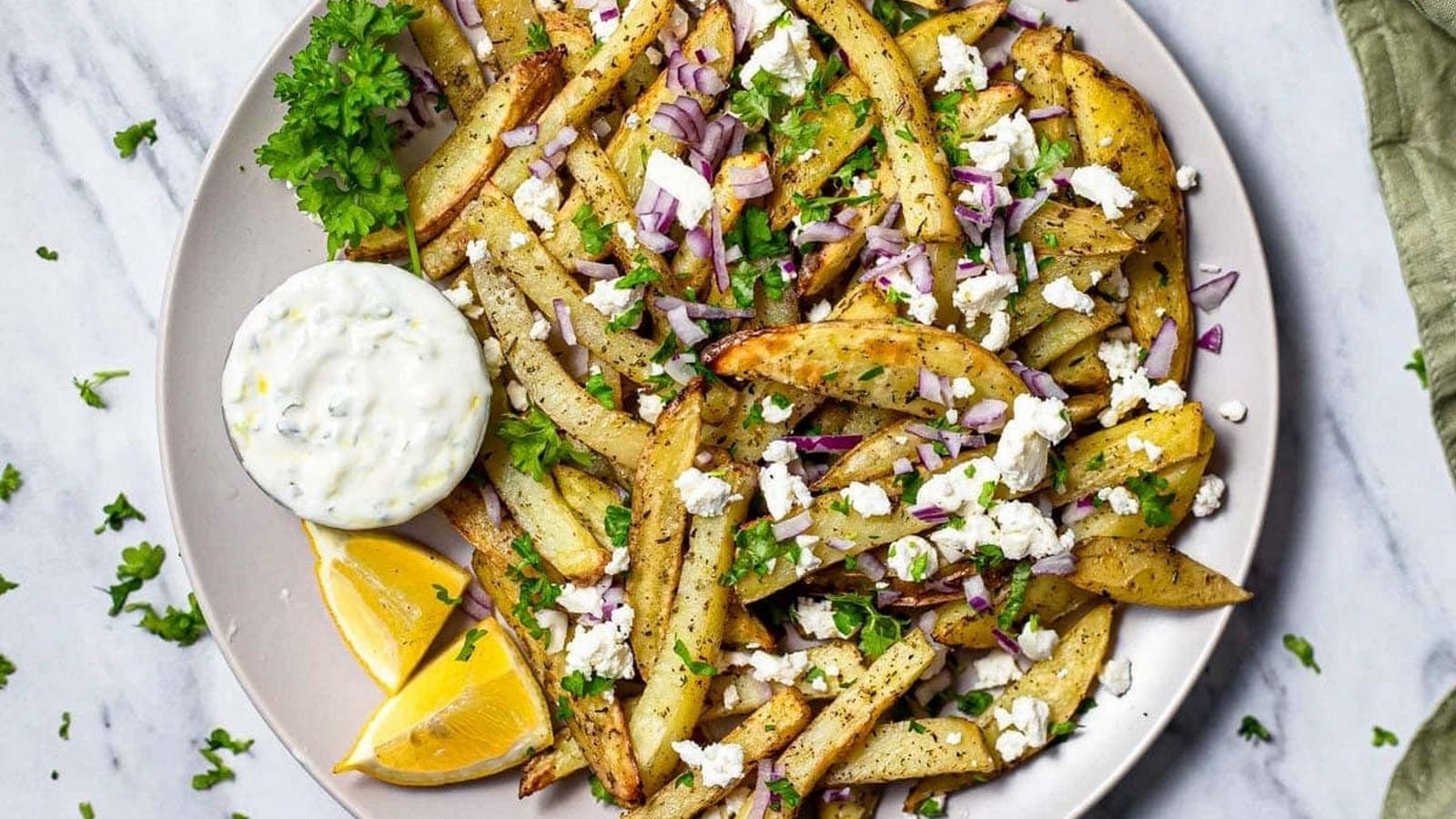 Plate of seasoned fries topped with feta cheese and red onions, garnished with parsley, a side of dipping sauce, and lemon wedges.