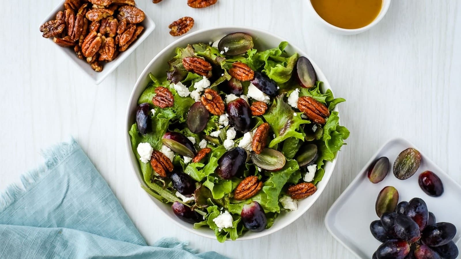 A bowl of salad with lettuce, grapes, pecans, and feta cheese. Nearby are small dishes of pecans and grapes, with a cup of dressing on the side. A blue napkin is on the table.