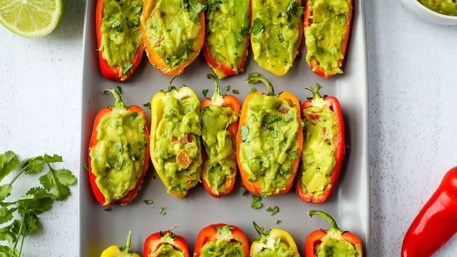 Mini bell peppers filled with guacamole are arranged neatly on a gray plate, garnished with fresh herbs.