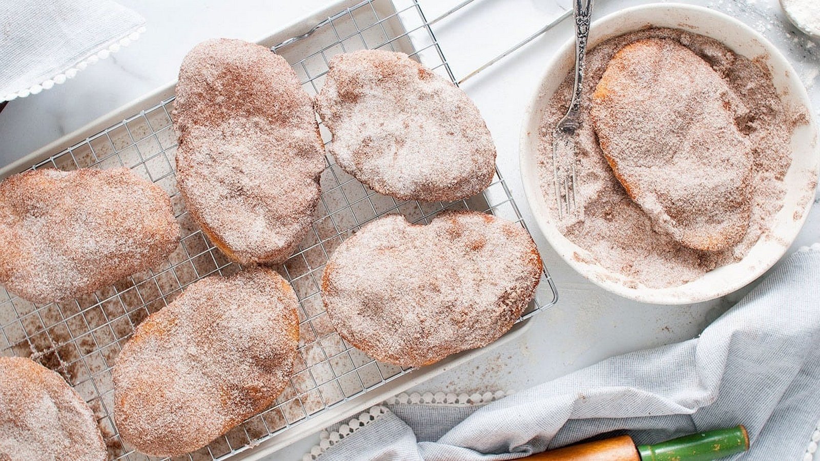 A rack of sugar-coated pastries next to a bowl of cinnamon sugar, with a pastry on a fork.