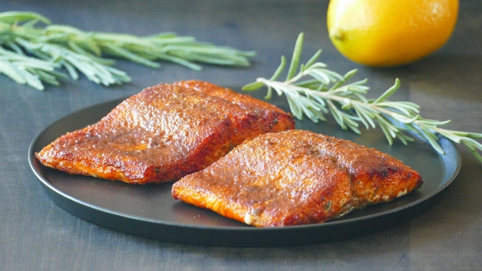 Two pieces of seasoned salmon on a black plate, surrounded by sprigs of rosemary, with a lemon in the background.