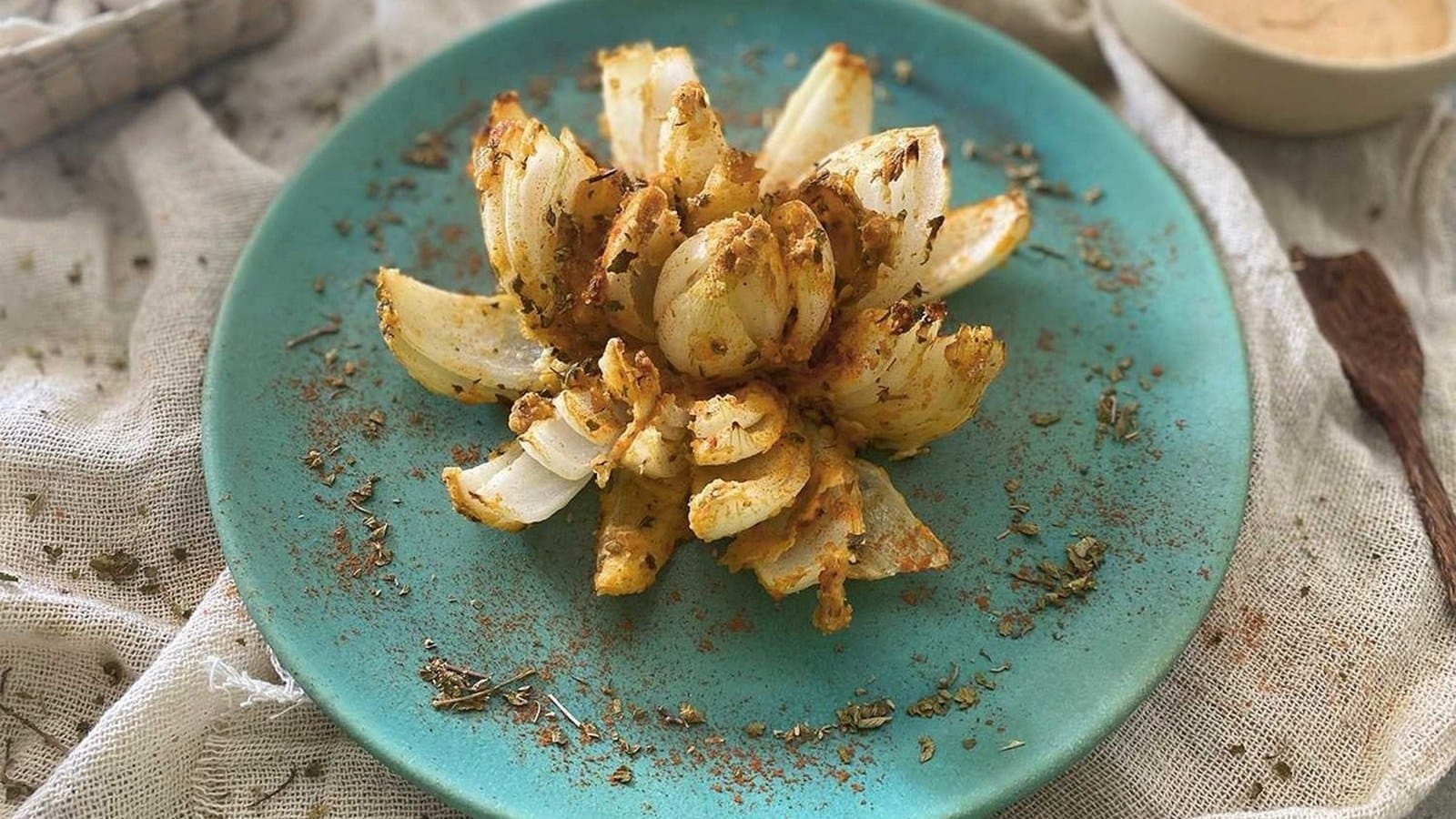 A crispy, fried blooming onion on a turquoise plate. The onion is seasoned and surrounded by scattered spices. A small dish of dipping sauce is in the background.