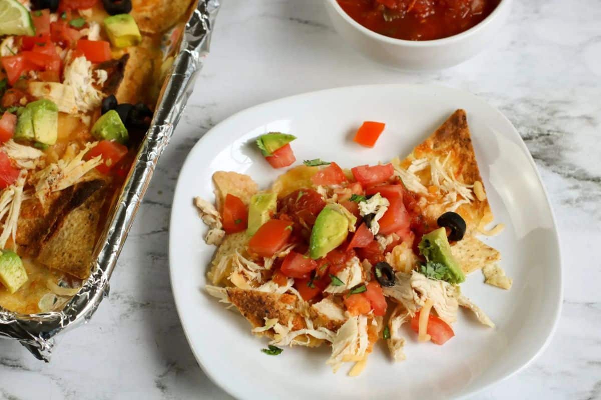 A white plate with nachos topped with shredded chicken, tomatoes, avocado, and olives. A tray of nachos and a bowl of salsa are in the background.