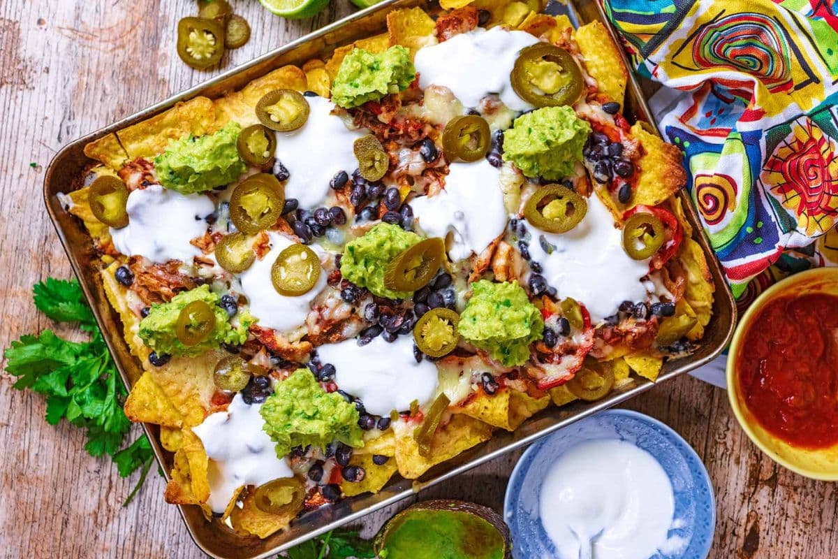 A tray of loaded nachos topped with guacamole, sour cream, jalapeños, black beans, and melted cheese, surrounded by colorful napkins and lime slices.