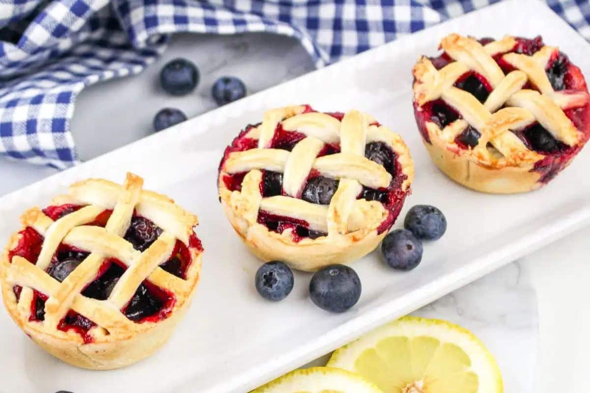 Three mini blueberry pies with lattice crusts on a white rectangular plate, surrounded by fresh blueberries and lemon slices, with a blue-checked cloth in the background.