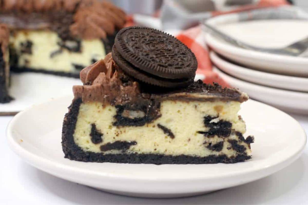 A slice of cookies and cream cheesecake on a white plate, topped with chocolate frosting and a whole chocolate sandwich cookie.