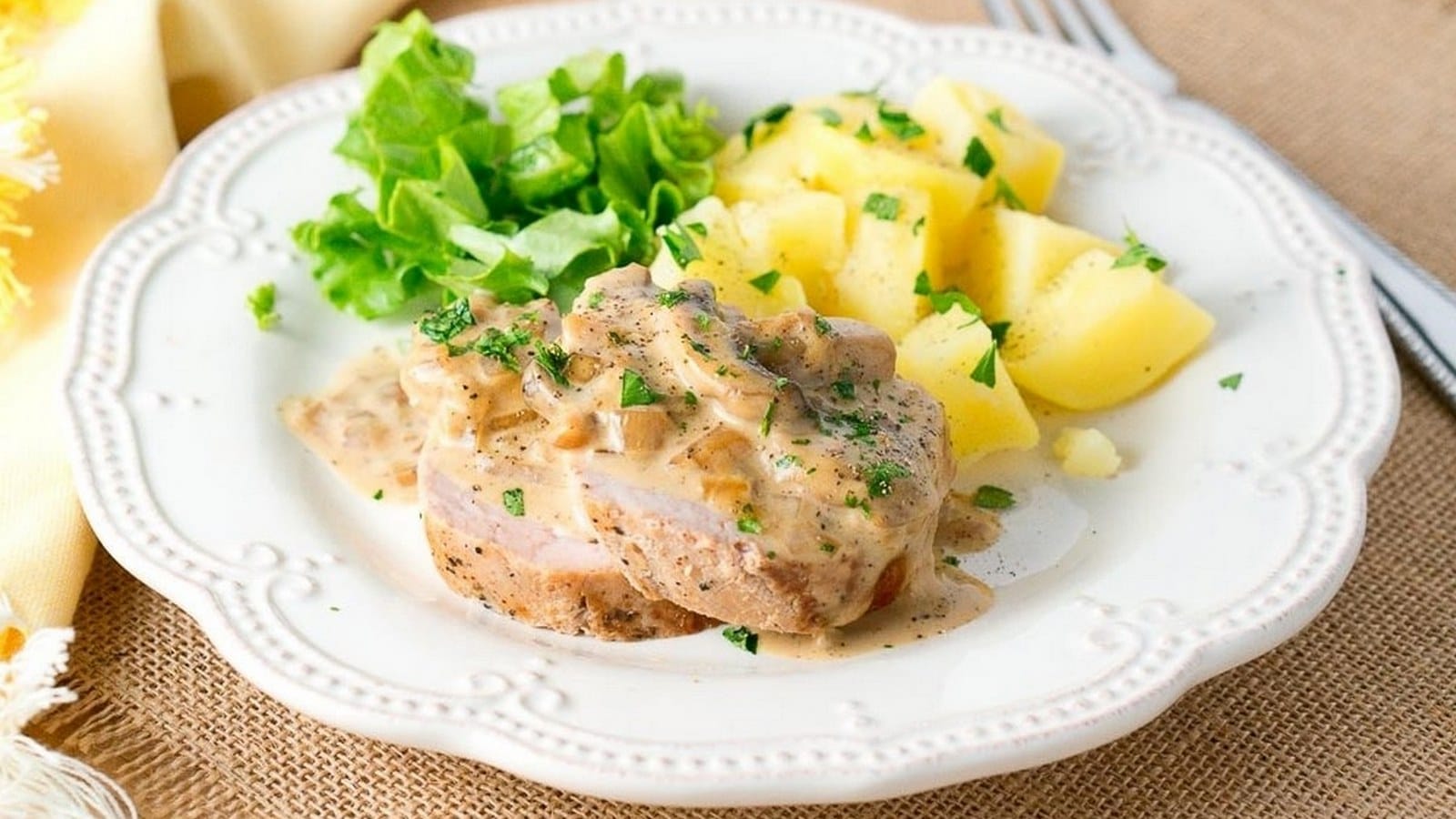 Plate with sliced pork covered in creamy mushroom sauce, accompanied by boiled potatoes and leafy greens on a white decorative plate.