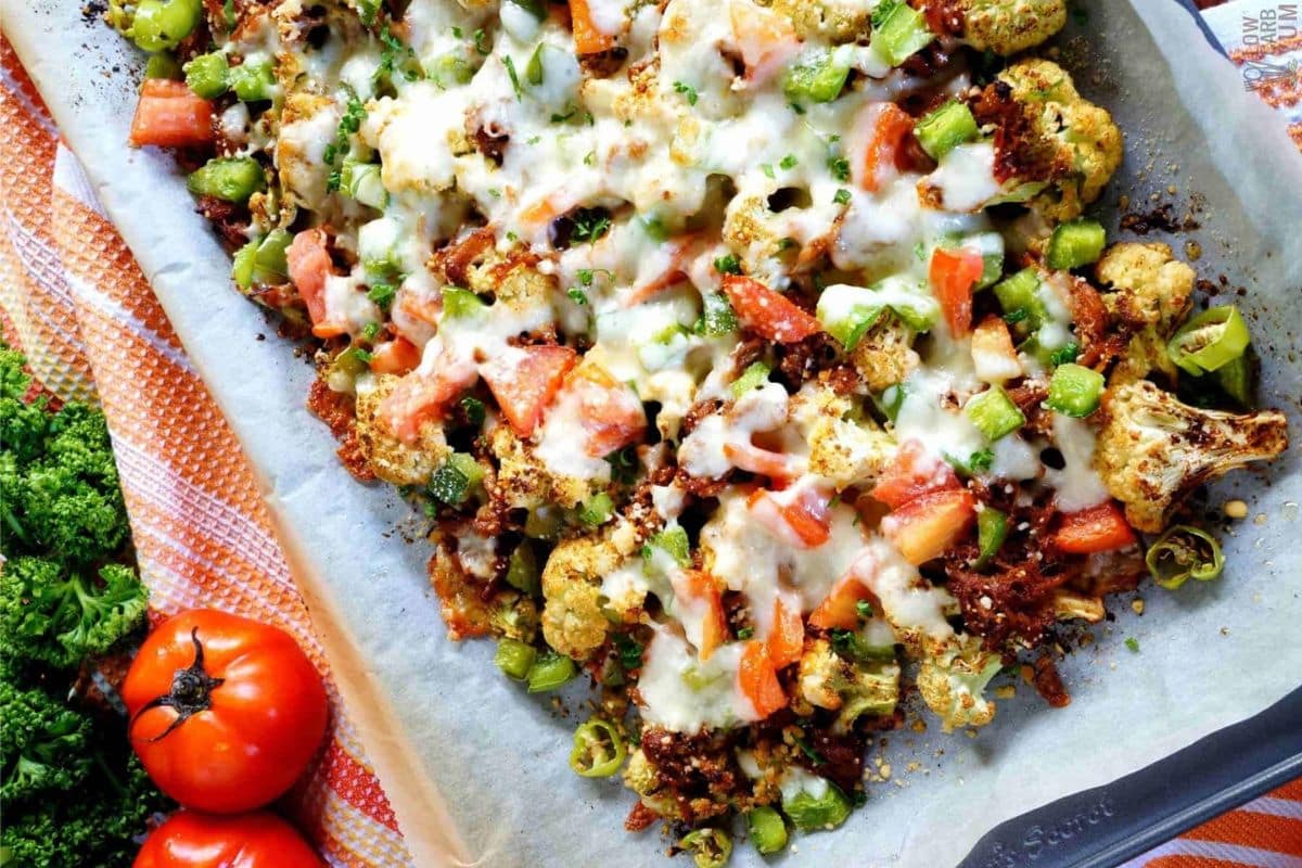 Baked dish with cauliflower, diced tomatoes, green peppers, melted cheese, and seasoning on parchment paper. Two whole tomatoes and parsley are nearby.