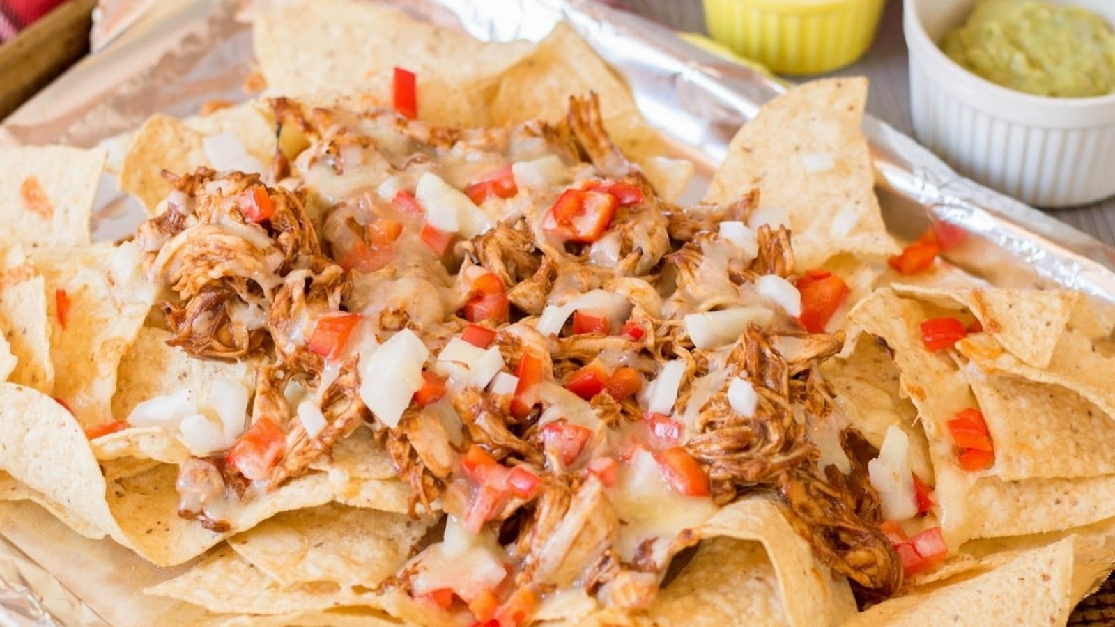 A tray of nachos with shredded chicken, melted cheese, diced tomatoes, and onions, served with guacamole in a small dish.