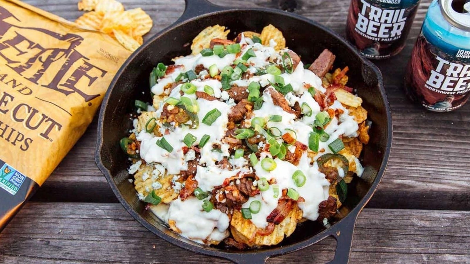 A cast iron skillet filled with loaded nachos topped with cheese, green onions, jalapeños, and sour cream. A bag of kettle chips and two cans of Trail Beer are nearby on a wooden table.