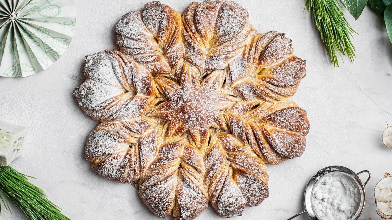 A star-shaped bread with layered petals, dusted with powdered sugar, placed on a light surface. Greenery and a strainer with sugar nearby.