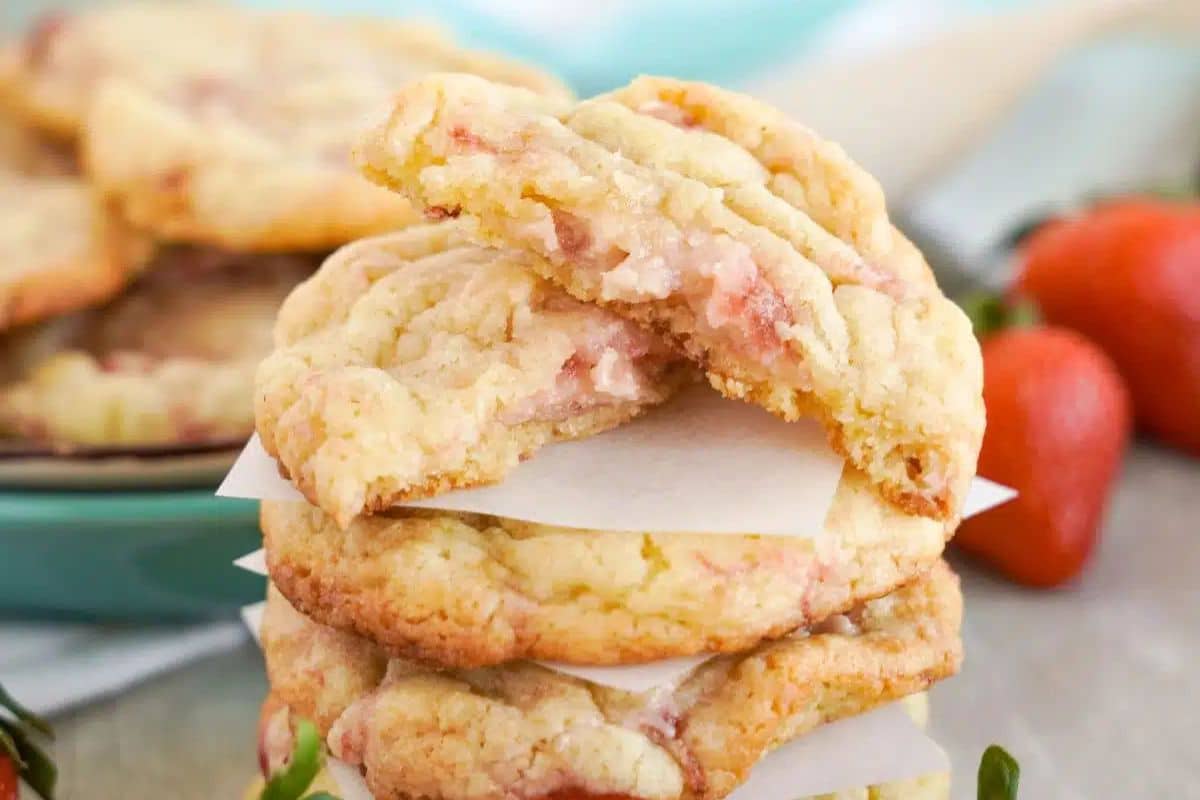 Stack of strawberry cookies with a bite taken from the top cookie, layered with parchment paper. Fresh strawberries are visible in the background.